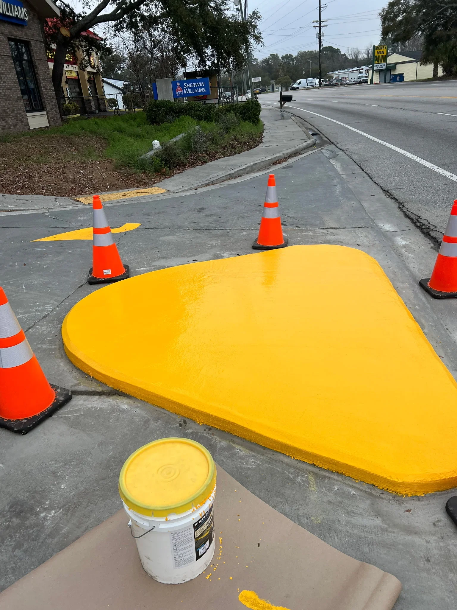 Yellow paint applied to asphalt parking lot with traffic cones and paint bucket visible